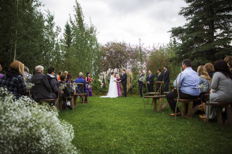 Bride and groom stand together holding hands while audience watches in grassy field surrounded by trees 
