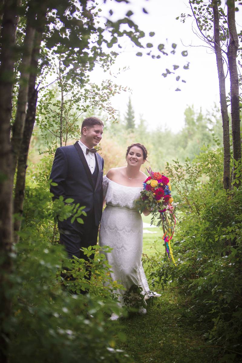 Bride and groom walk down grassy forest pathway holding multicolored bouquet 