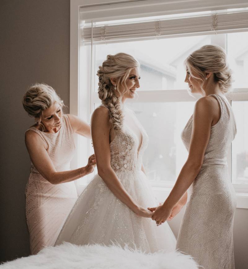 Bride holds hands of woman while mother adjusts dress white lace dress from behind 