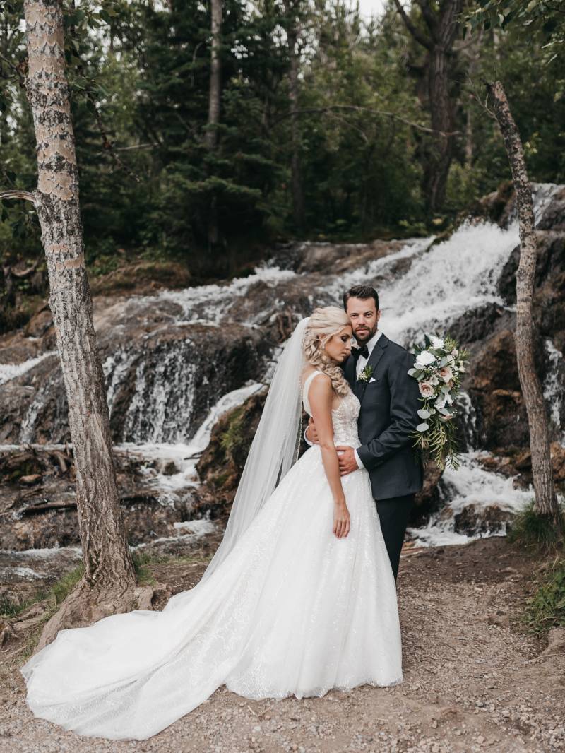 Bride stands embraced by groom in front of woodland river and waterfall 