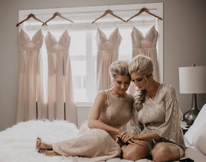Two women sit in white bed in front of four hanging wedding dresses on window frame 