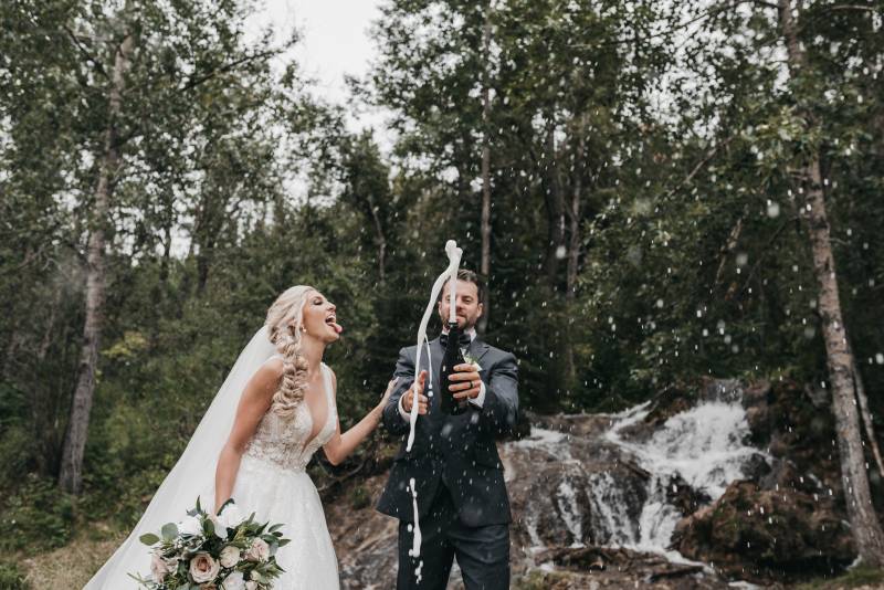 Groom holds foaming bottle while bride stands tongue out in front of woodland waterfall 