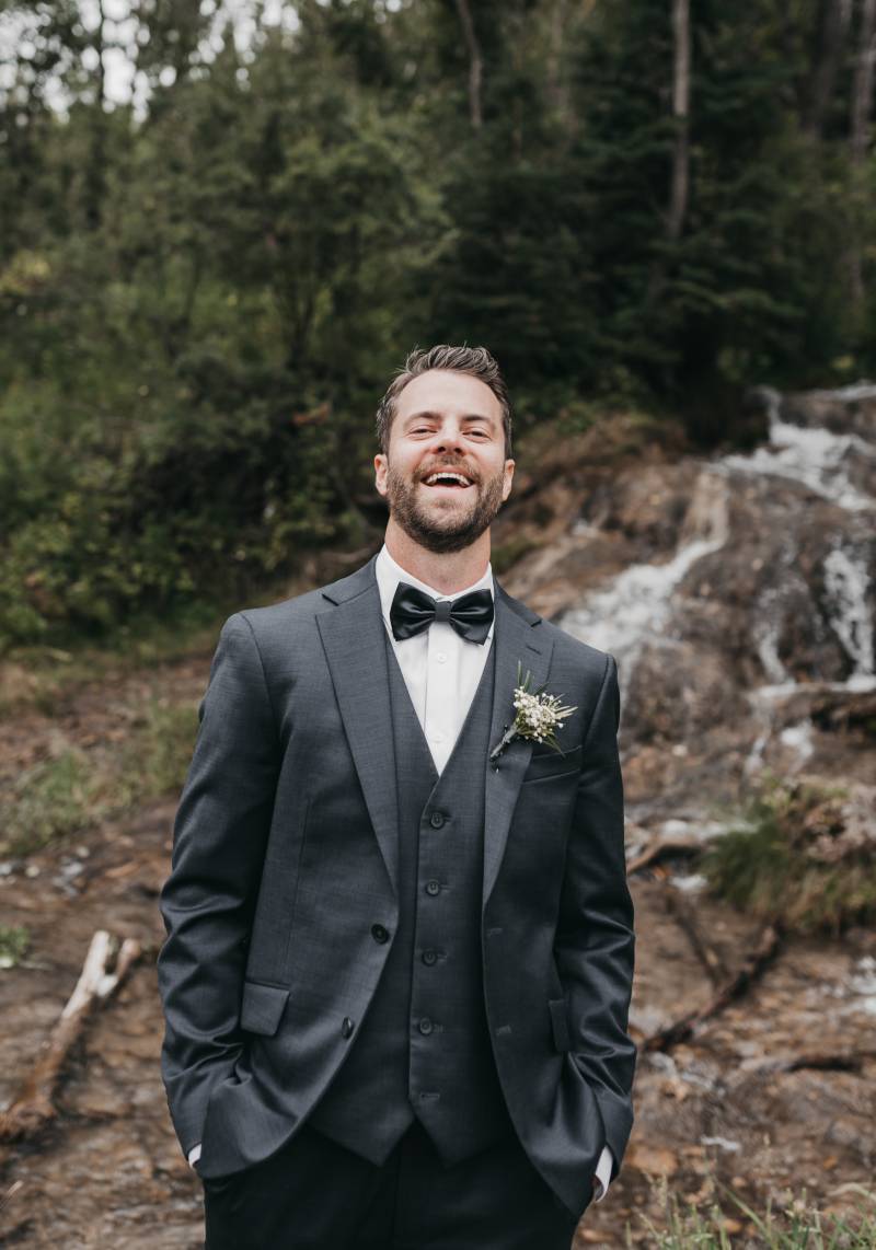 Groom smiles hands in pockets of black suit standing in front of  small woodland waterfall 
