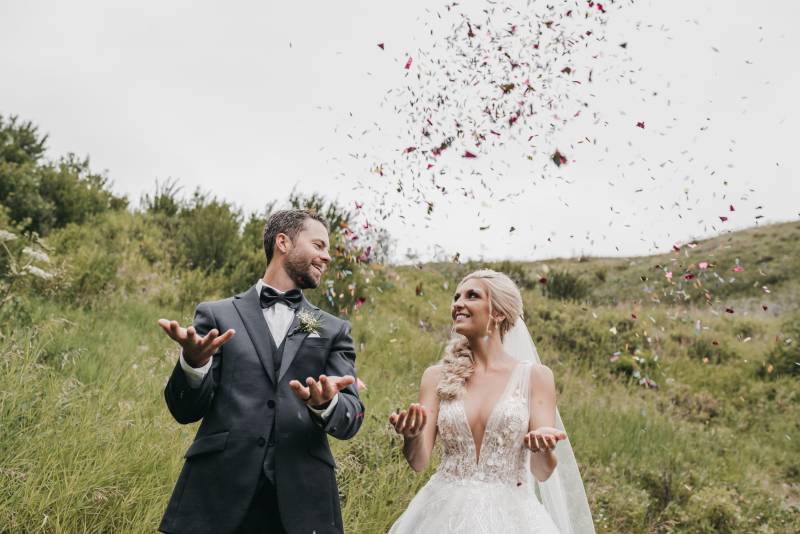 Bride and groom throw petals up hands out smiling in grassy hills 
