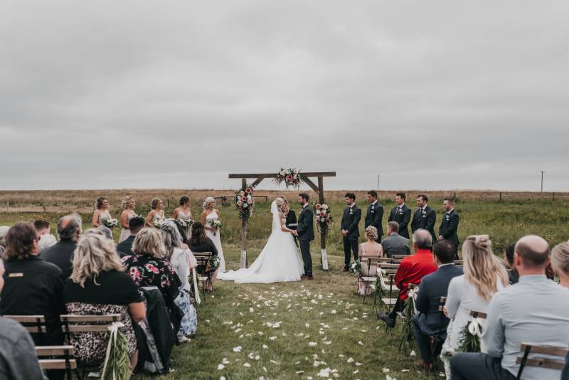 Bride and groom holds hands under wooden wedding arch while guests sit petals on ground 
