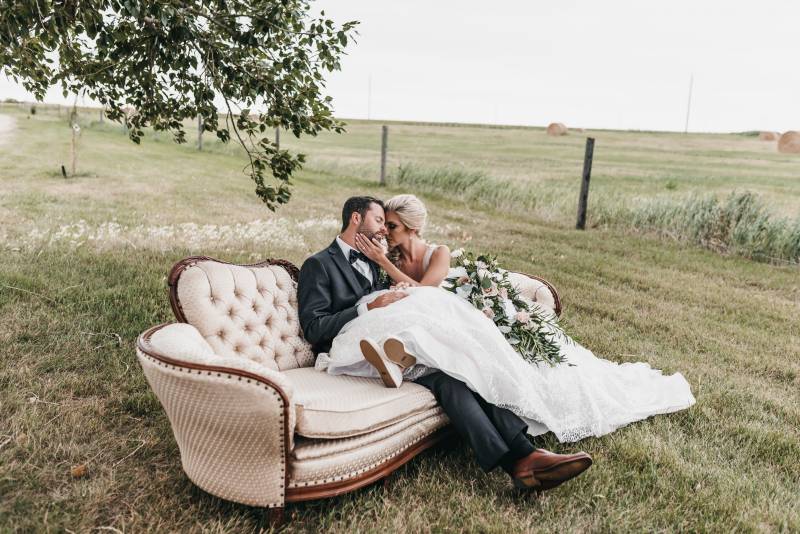 Bride sits across grooms lap sitting on sofa in the middle of grassy field 