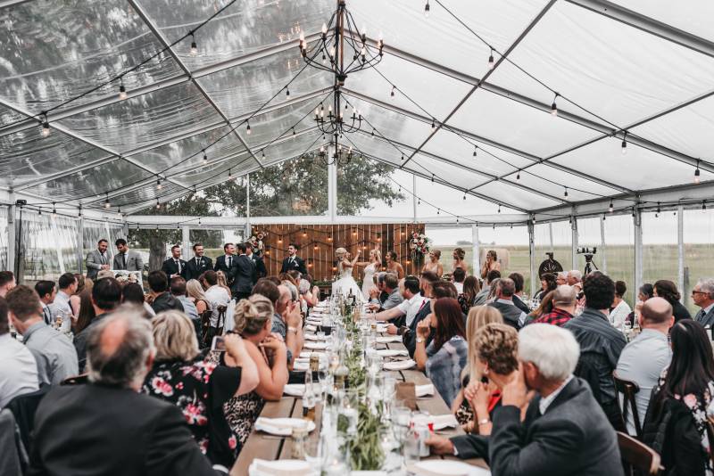 Wedding reception under clear tent with hanging lights and chandeliers guests seated 