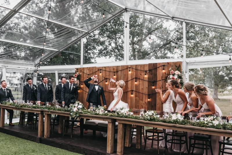Bride and groom stand behind head table drinking bottle as bridesmaids and groomsmen watch