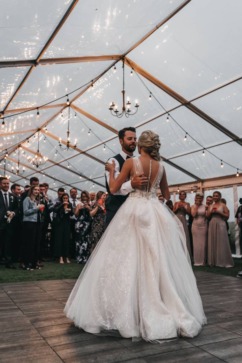Bride and groom dancing under hanging lights while guests stand behind 