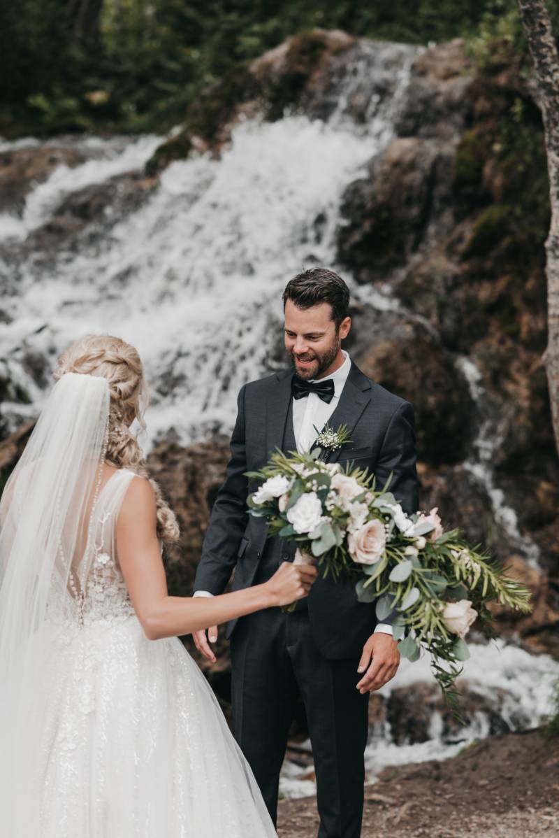Bride walks towards groom wearing white lace dress and veil holding blush and white bouquet in front of waterfall 