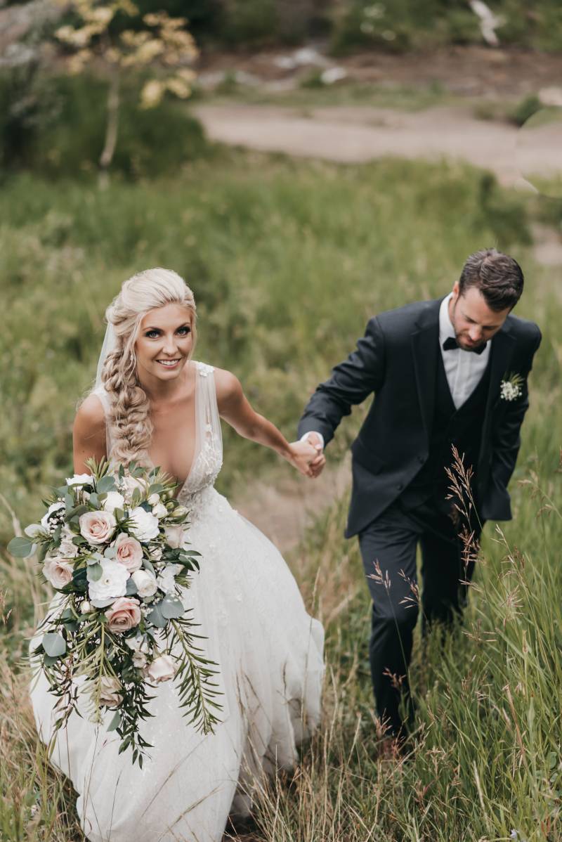 Bride smiling leading groom up grassy hill holding blush and white bouquet 