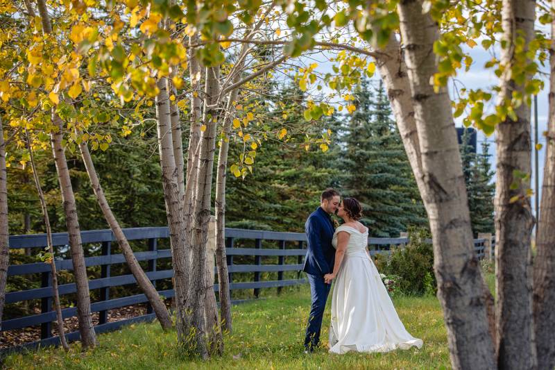 Bride and groom stand close holding hands under yellow green fall leaves 