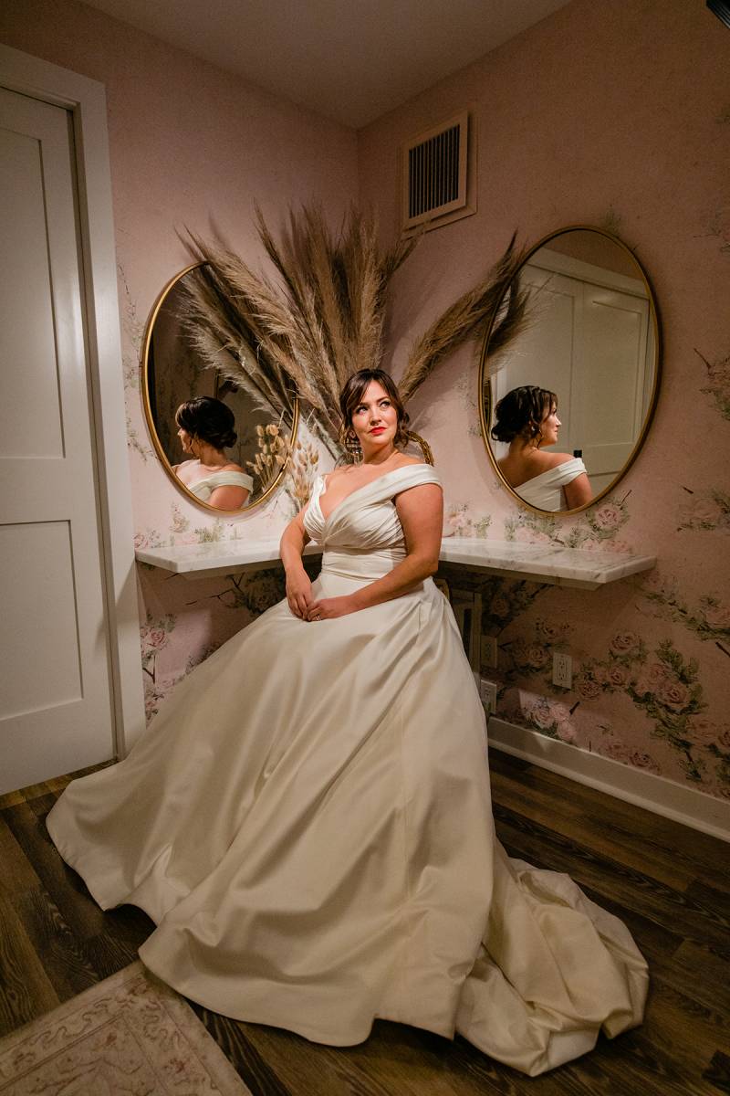 Bride sits in large white dress looking to side in front of mirrors and pampas grass 