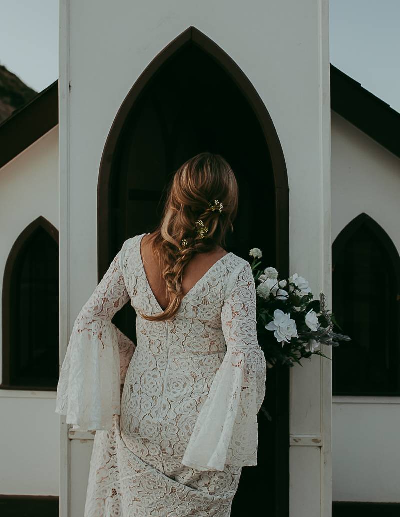 Bride stands in white doorway in white lace dress holding white bouquet 