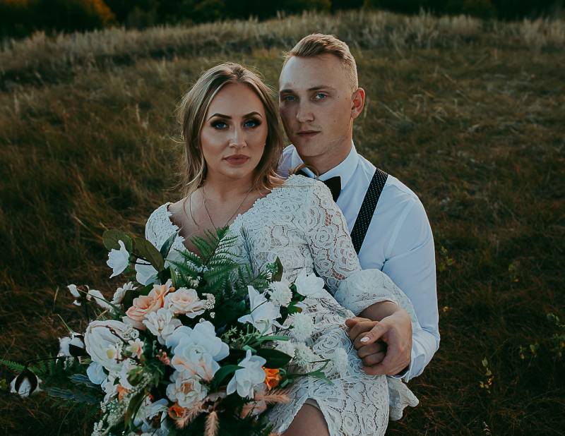 woman sits in mans lap on grassy hill holding peach and white bouquet 