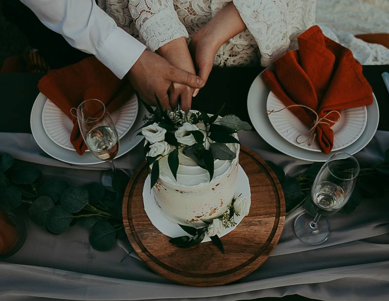 Man and woman cut white wedding cake together beside place settings with orange napkins