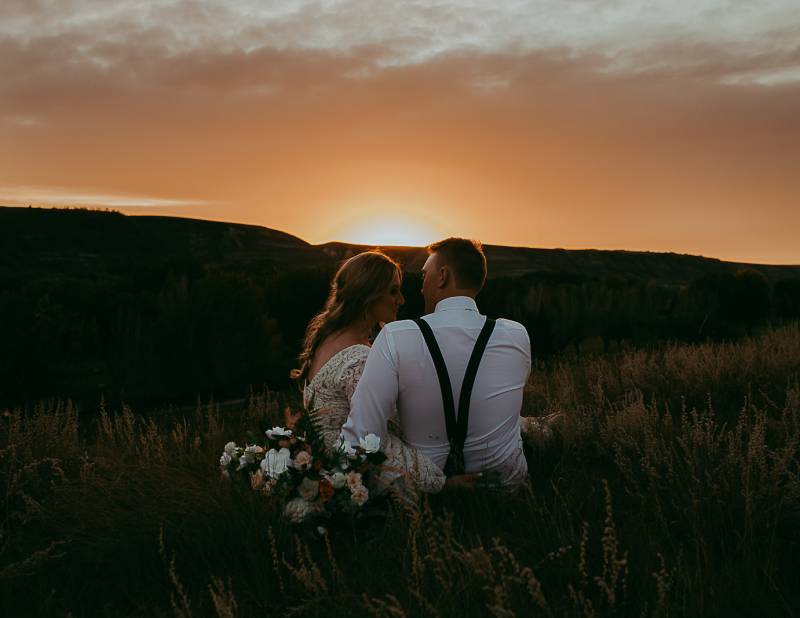 Man and woman sit in grassy field bouquet behind at sunset 