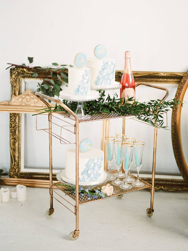 Golden trolley with three white and powder blue wedding cakes beside pink bottle and gold rimmed glasses 