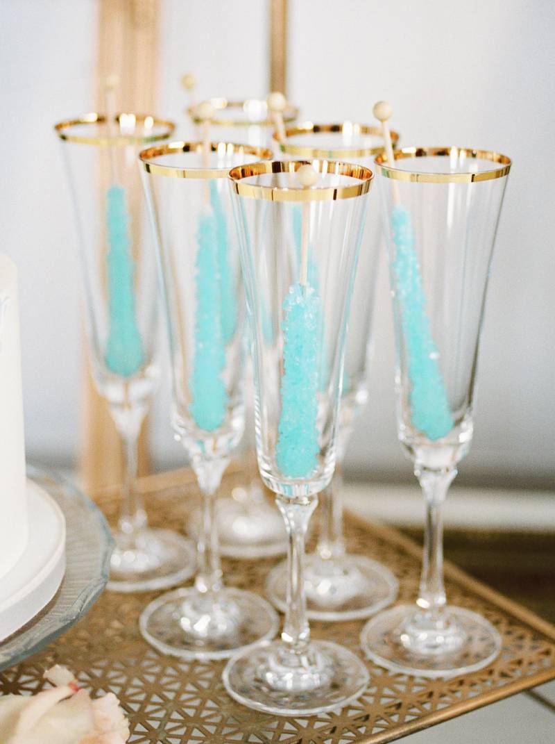 Six tall glasses with golden rim and light blue rock candy inside on gold shelf
