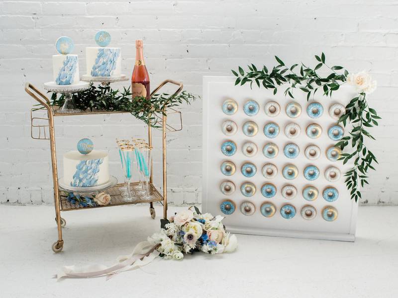 Gold Trolley with white and blue cakes and rose bottle beside mini doughnut wall and white bouquet 