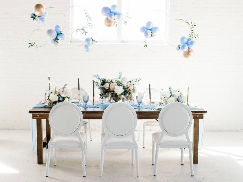 Dark brown wood table and white chairs with powder blue table setting and table runner with hanging blue and gold balloons
