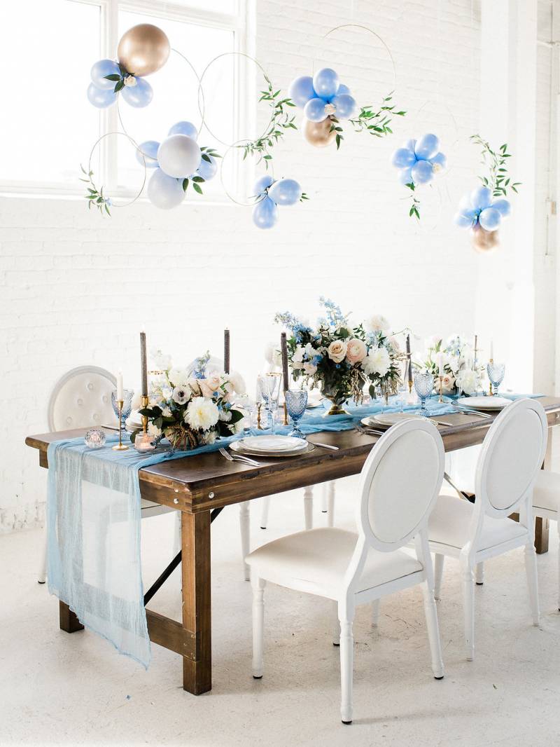 Dark brown wood table and white chairs with powder blue table setting and table runner with hanging blue and gold balloons