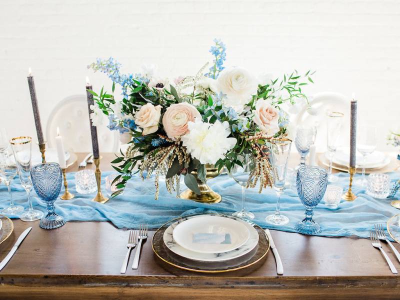 Dark brown wood table with powder blue table setting and table runner holding gold potted floral centerpiece 