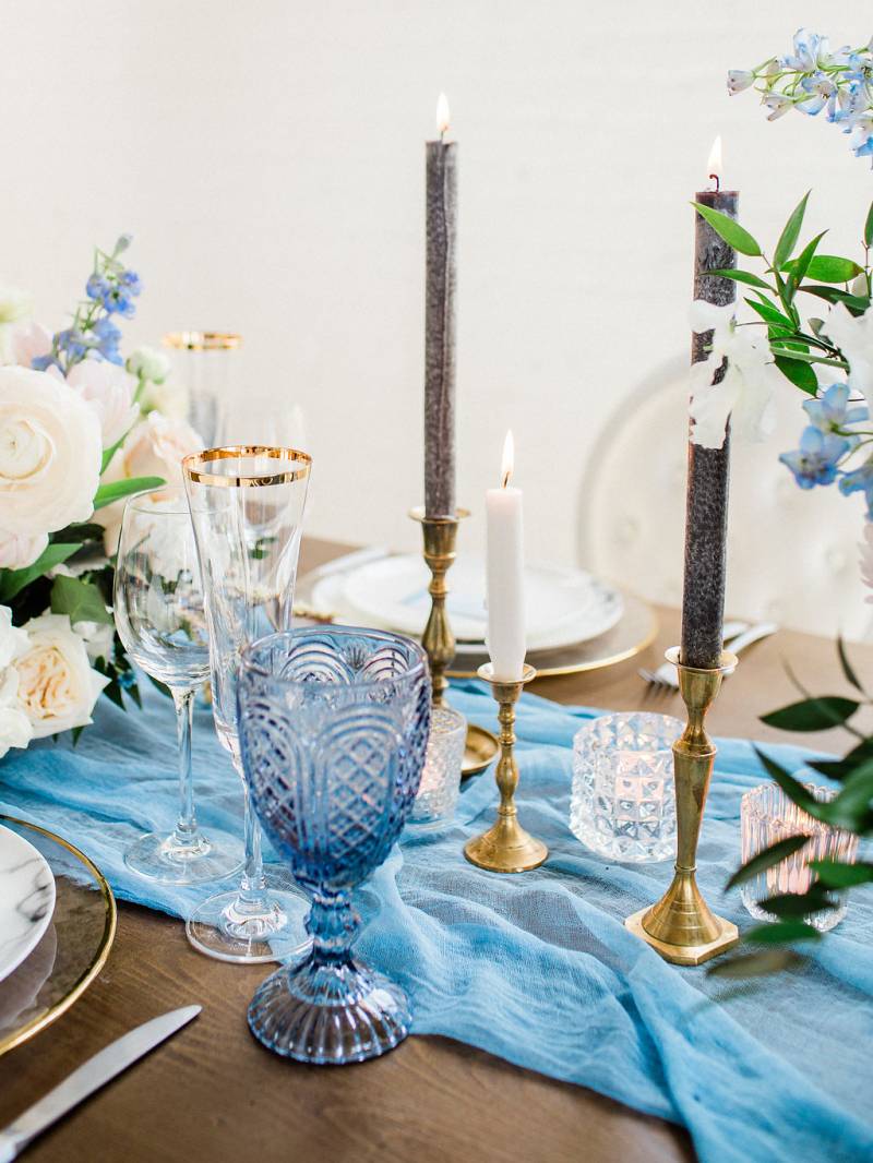 Dark brown wood table with powder blue table setting holding gold potted floral centerpiece and gold taper candles