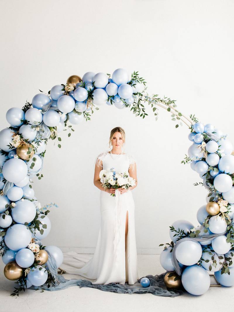 Woman in white dress with slit stands under powder blue balloon wedding arch