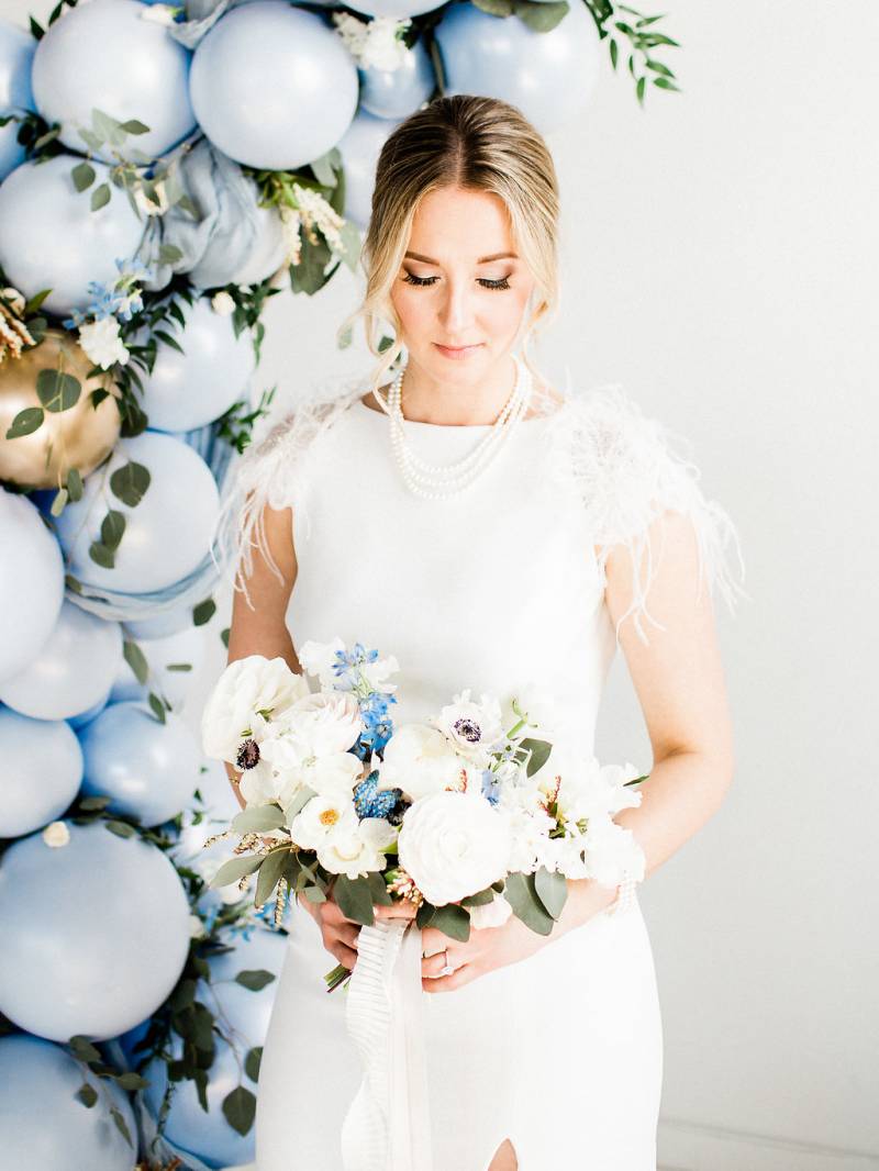 Woman in white dress stands beside powered blue balloon arch holding bouquet 