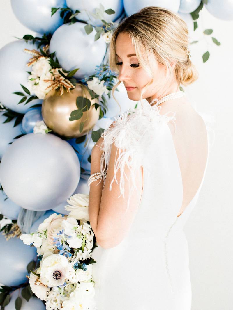 Woman in white open back dress stands facing powered blue balloon arch holding bouquet 