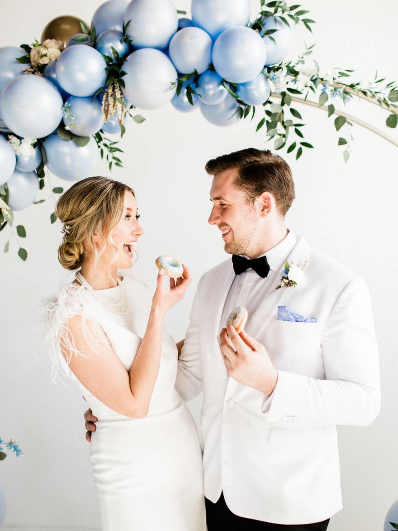 Man and woman smile under powder blue wedding arch with gold accents holding blue frosted mini doughnuts