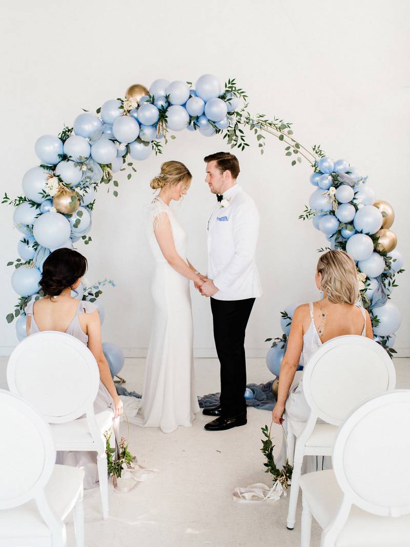 Man and woman hold hands under powered blue balloon wedding arch guests watch 