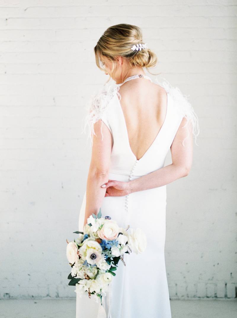 Woman in white open back dress facing wall with white bouquet in hand 