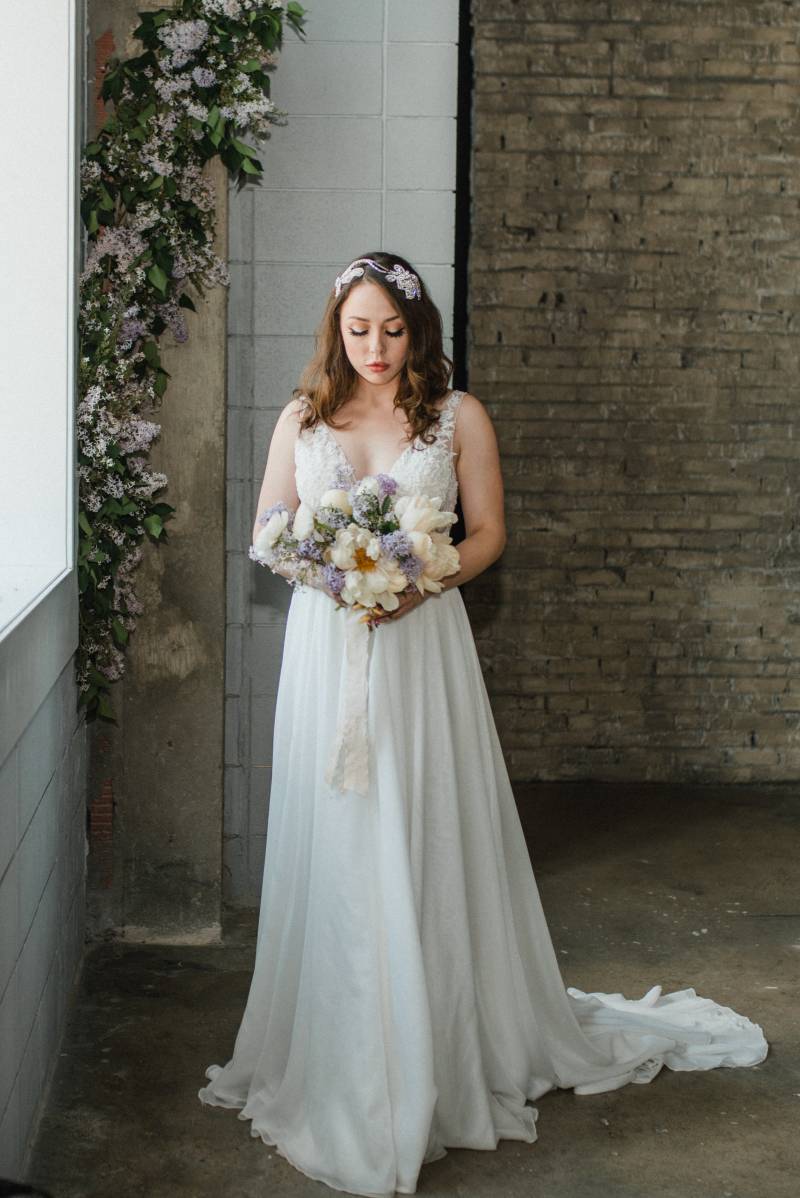 Woman in white lace dress holding white and lilac bouquet beside window with lilac accents 
