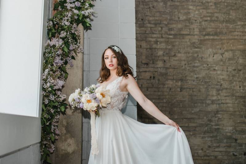 Woman in white lace dress holding white and lilac bouquet under lilac arrangement 