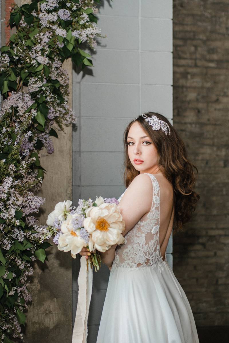 Woman in white lace dress looking over shoulder holding white and lilac bouquet 