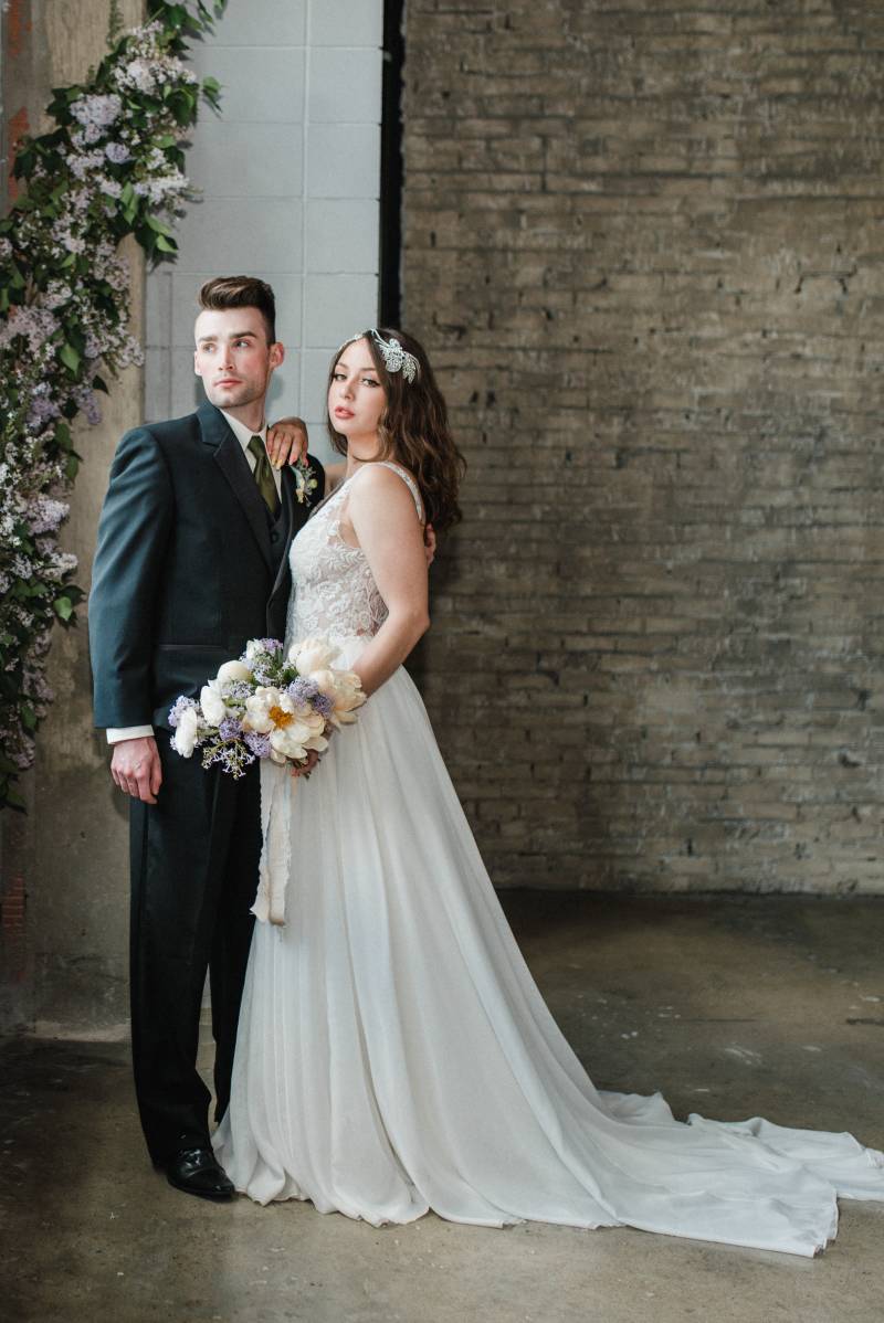 Woman in white lace dress leans on shoulder of man in black suit looking into distance 