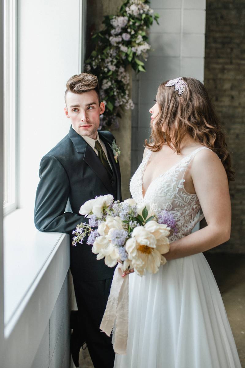 Woman holding white and lilac bouquet looks over shoulder at man leaning on window sill in black suit 