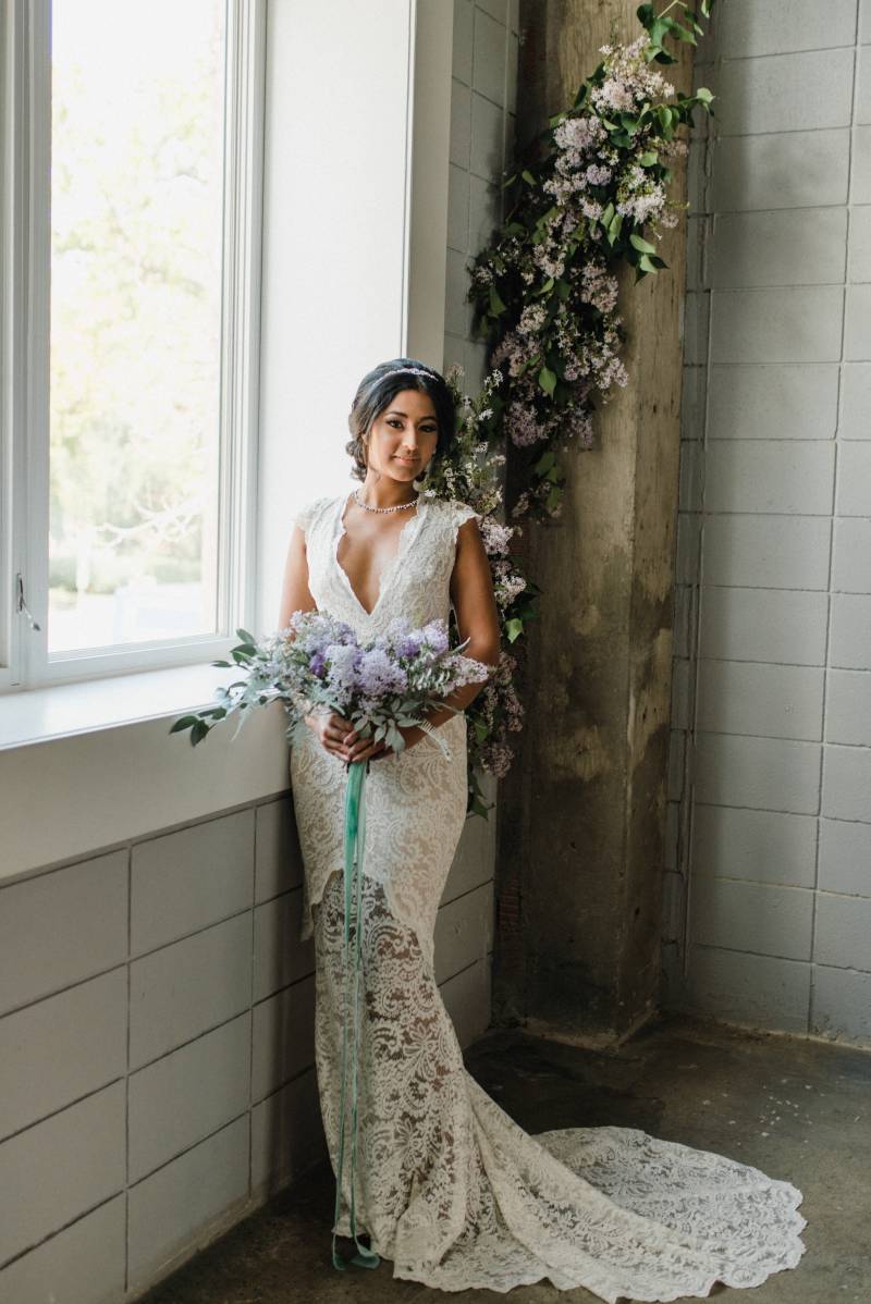 Woman in white lace dress stands leaning against window sill holding lilac bouquet 