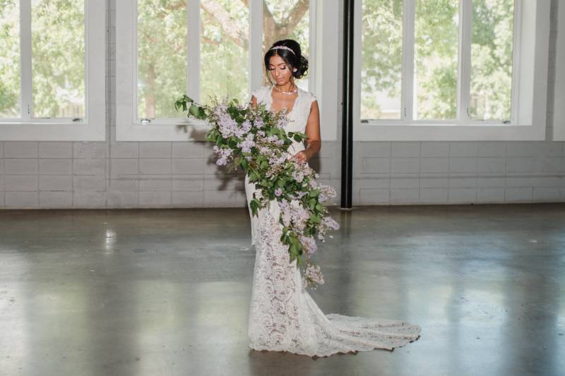 Woman in white lace dress stands in open room holding large lilac floral arrangement 
