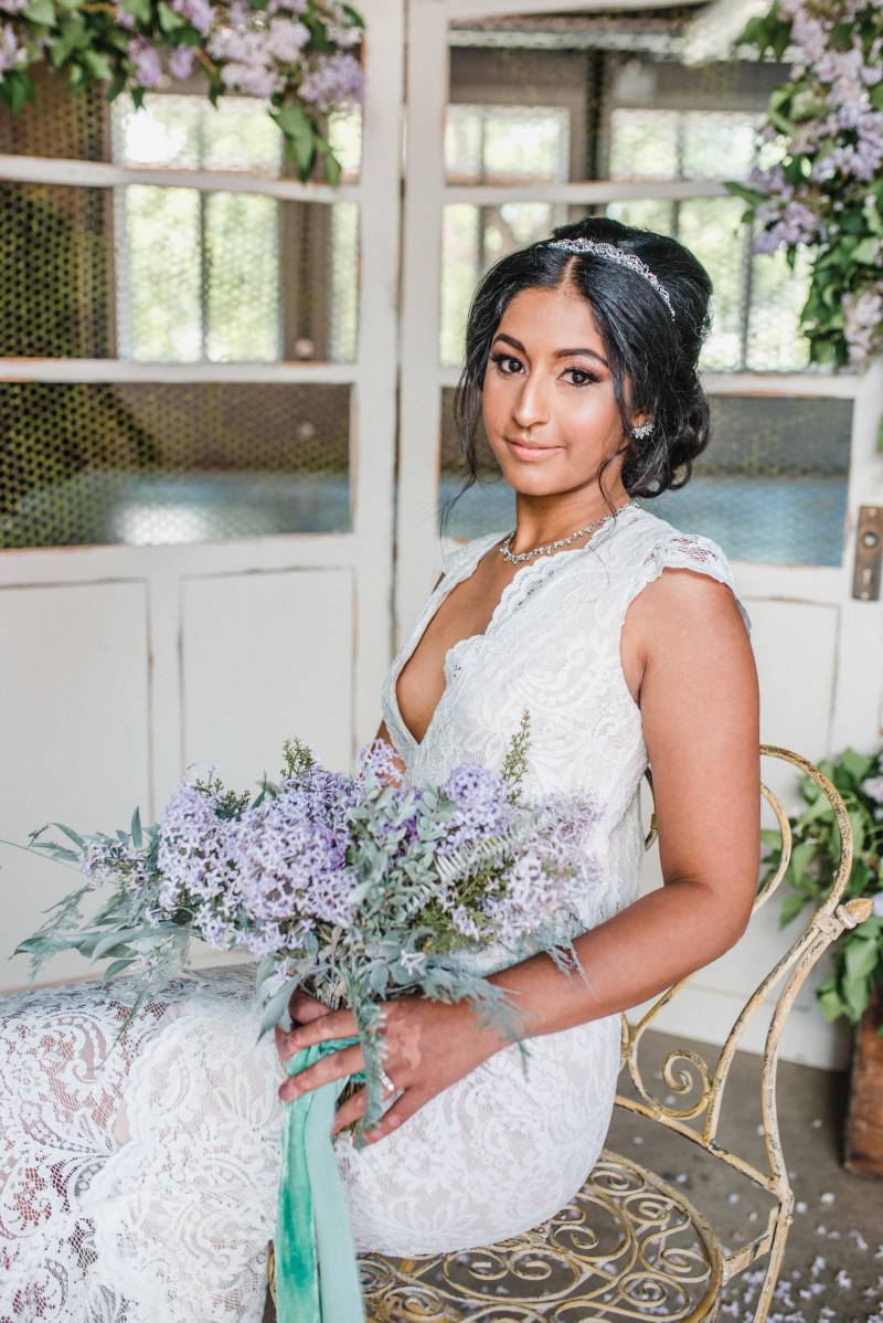 Woman in white lace dress sits holding lilac bouquet in front of white doors detailed with lilac accents 