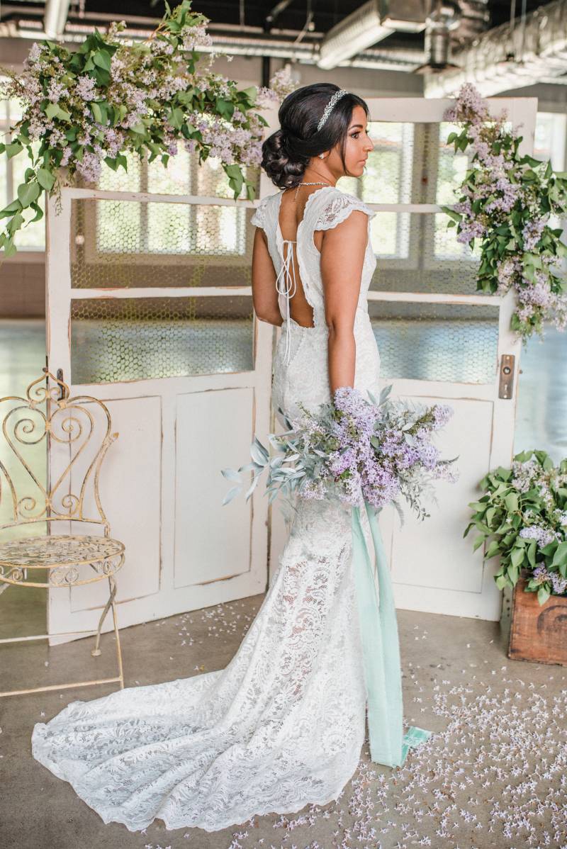 Woman in white lace dress holding lilac bouquet to side in front of lilac detailed white doors 