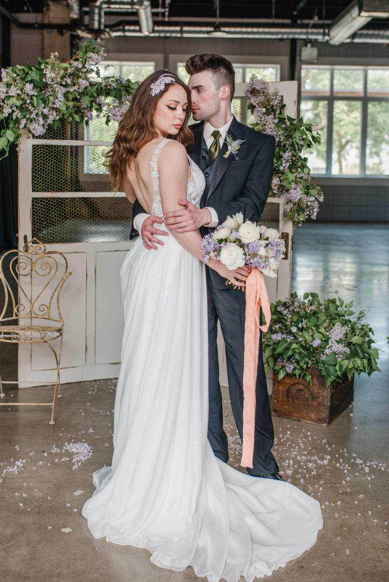 Man and woman embrace in front of white doors with lilac accents and yellow metal chair 