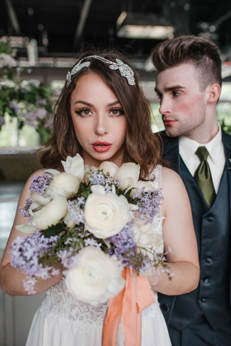 Woman in white lace dress holding white and lilac bouquet man looks to side behind 