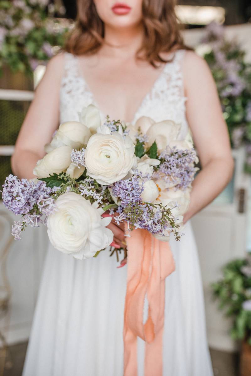 Woman in white lace dress holding white and lilac bouquet with peach ribbon 