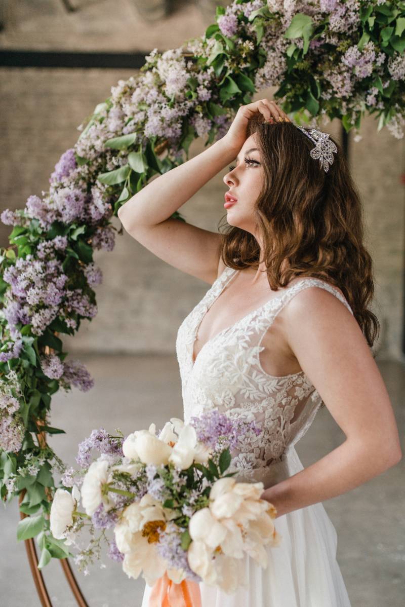 Woman in white lace dress holding white and peach bouquet touching hair under copper wedding arch with lilac accents 