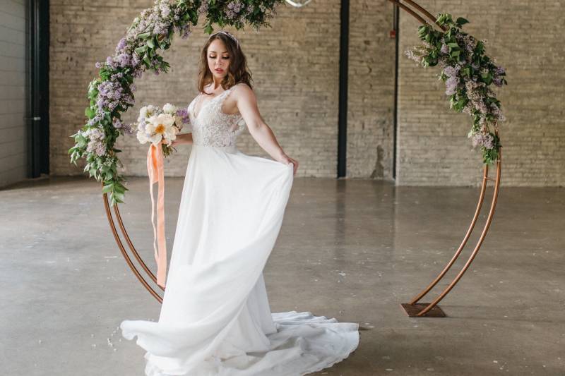 Woman lifts skirt to side holding white and peach bouquet under copper wedding arch with lilac accents 
