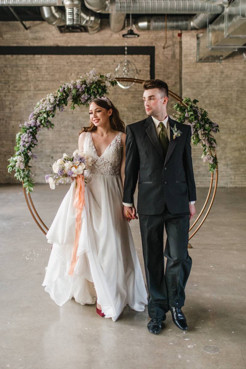 Man and woman walk away holding hands from copper wedding arch and lilac accents 