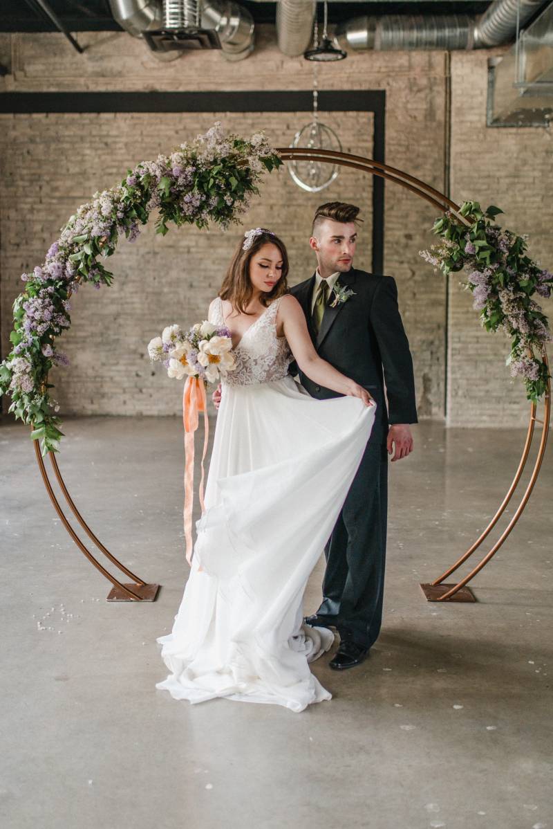 Man and woman stand as woman lifts skirt to side in front of copper wedding arch and lilac accents 